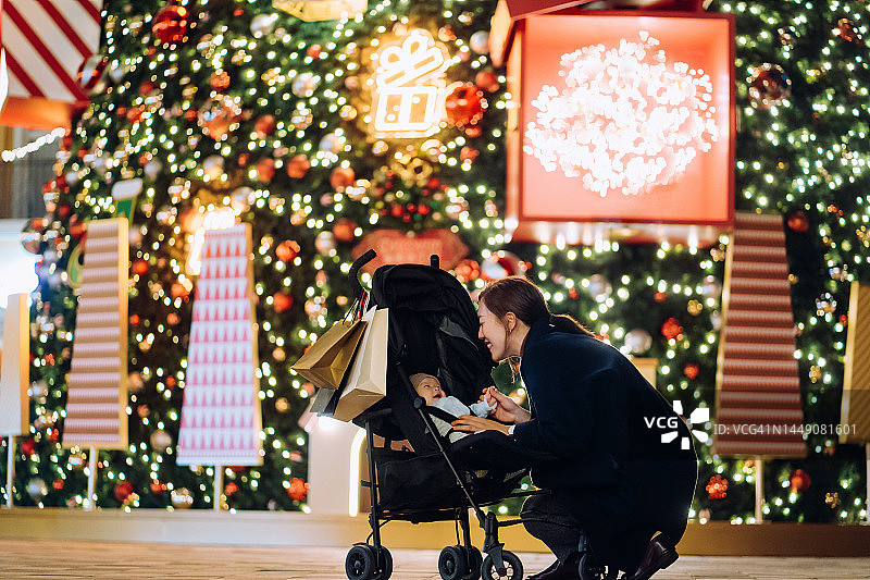 充满爱意的年轻亚洲母亲在圣诞市场与她可爱的女儿在婴儿车里玩耍，一起快乐地微笑。背景是灯火通明的圣诞树。家庭温馨时刻。节日气氛。圣诞节来了图片素材