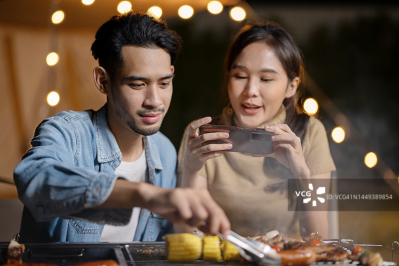 科技让生活更快乐：亚洲男人在户外露营烧烤腌制牛肉，朋友拍照分享图片素材