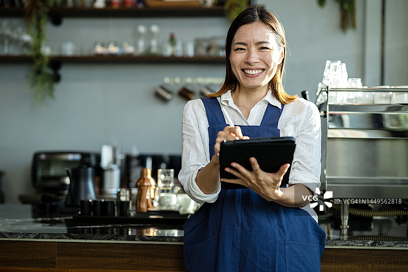 通过咖啡店特许经营赚钱：日本咖啡店女店主站在收银台前，手持平板电脑接收顾客订单图片素材