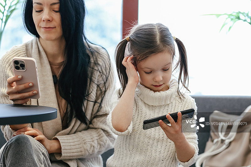 在城市咖啡馆使用手机的可爱5岁女孩和她的年轻黑发母亲图片素材