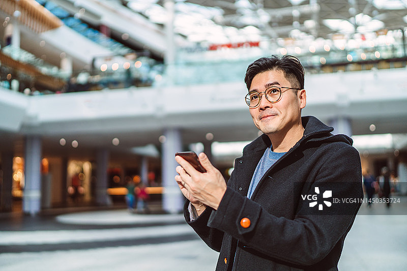 年轻的亚洲男人在购物中心高兴地使用智能手机图片素材