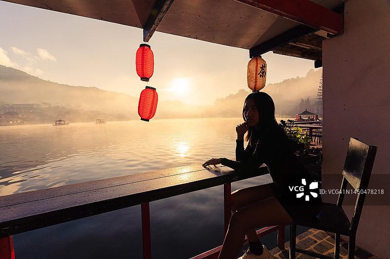 一个女人坐在雾气蒙蒙的河边，村庄和山脉，阳光普照。Ban Rak Thai，泰国Mae Hong Son。图片素材