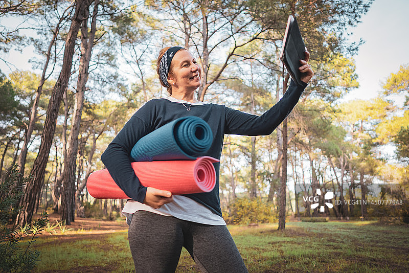 年长的女人手拿瑜伽垫在森林里自拍微笑图片素材