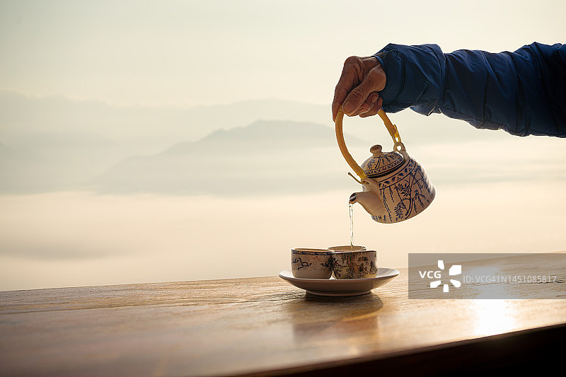 老人的手将中国茶倒入白色杯中，背景是雾蒙蒙的山间日出图片素材