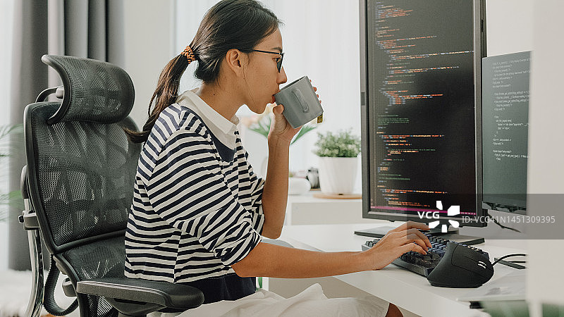 在家远程工作，喝咖啡并使用电脑编写代码的年轻亚洲女性软件开发人员图片素材
