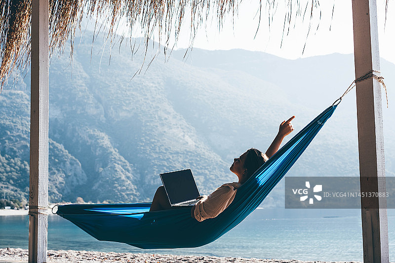 夏季休假兼顾工作和休闲的游客：在海滩吊床上使用笔记本电脑的女人图片素材