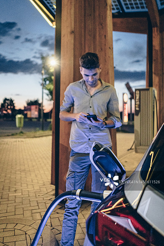 年轻男子在充电站为电动汽车充电，使用手机图片素材