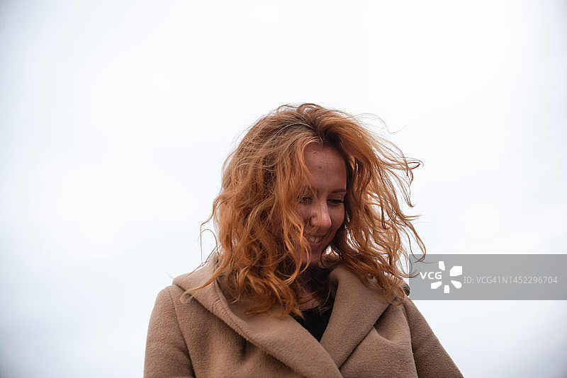 灰色天空下，身穿米色外套的微笑红发年轻女人像图片素材