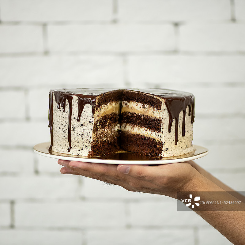 手中盘子里的切块蛋糕：家庭庆祝的奶油巧克力甜点，白色砖墙背景，柔焦，侧视图，剖视图图片素材