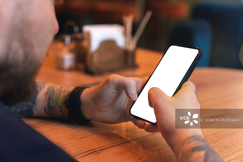 有纹身和胡须的男人手中拿着带复制空间的空白手机、肩后视角、网上冲浪、在线购物、远程工作概念图片素材