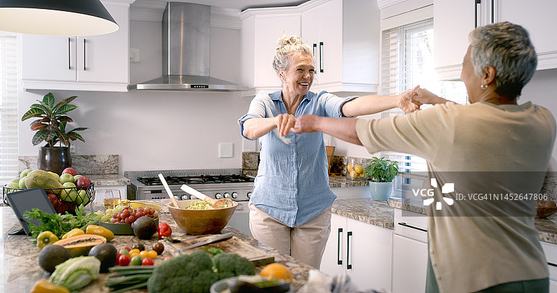 朋友们在厨房里跳舞、做饭，快乐地微笑，在家中一起享受多元化的乐趣音乐。女人们在家里准备饭菜、水果和蔬菜时感到幸福和欢快图片素材