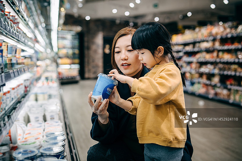 年轻的亚洲母亲和她的小女儿在超市购物，在乳制品区选择乳制品，阅读有机健康酸奶容器上的营养标签，健康饮食图片素材