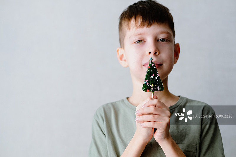 穿着绿色T恤的男孩在白色墙壁前吸吮圣诞树形状的棒棒糖图片素材