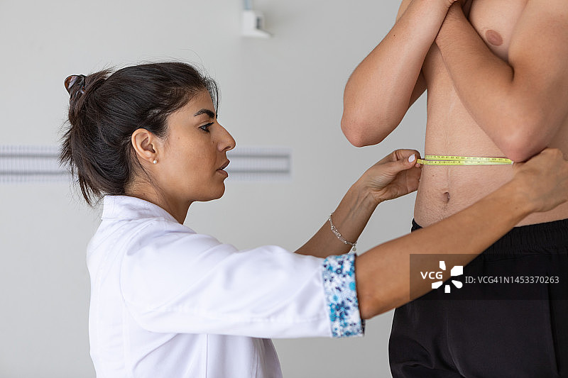 女营养师和体态专家在办公室进行医疗咨询，用卷尺测量青少年男病人的腰围图片素材
