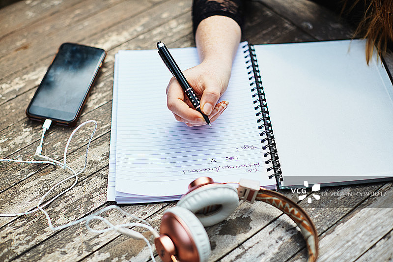 年轻女子在户外桌旁用螺旋笔记本写作，配有手机和耳机图片素材