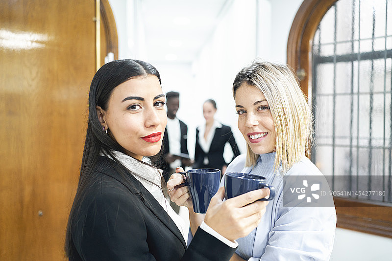 办公室走廊里身穿商务装、直视镜头的女同事，同事们在休息时间喝咖啡。图片素材