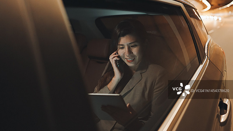 快乐的中国亚洲女商人坐在后排乘客座位上，在城市夜晚行驶时打电话图片素材