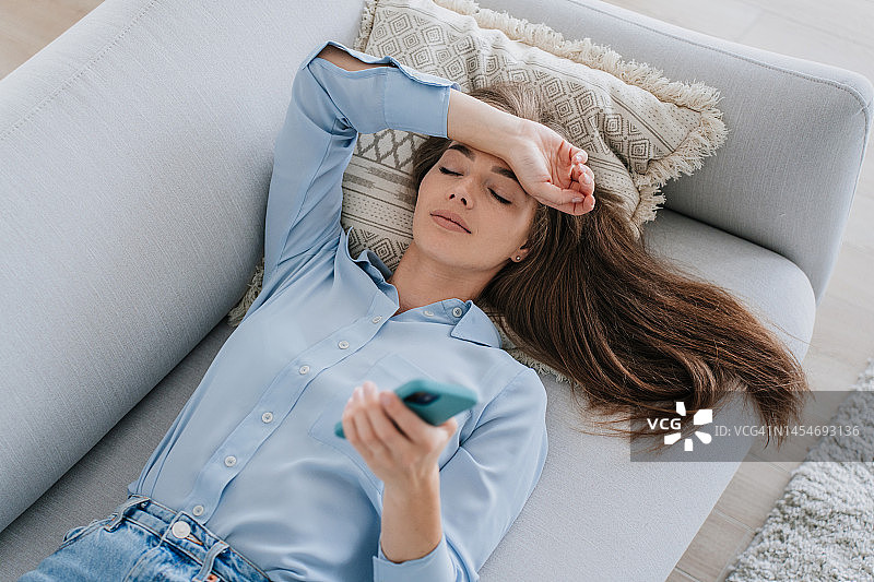 疲惫的年轻意大利女子身穿蓝色衬衫和牛仔裤躺在沙发上，闭着眼睛，手扶额头图片素材