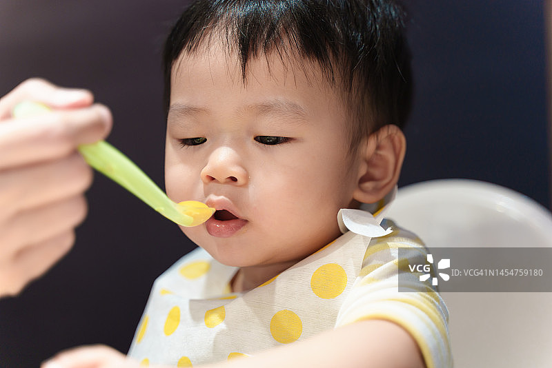 坐在高脚椅上快乐的亚洲可爱宝宝享受用餐，母亲用勺子喂可爱的宝宝，亚洲家庭家庭生活方式概念。图片素材