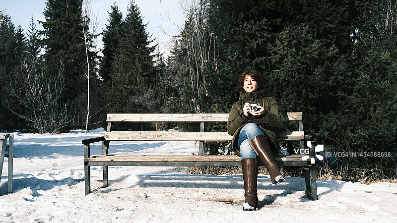 女人坐在阳光明媚的冬季公园长椅上，使用老式胶片相机图片素材
