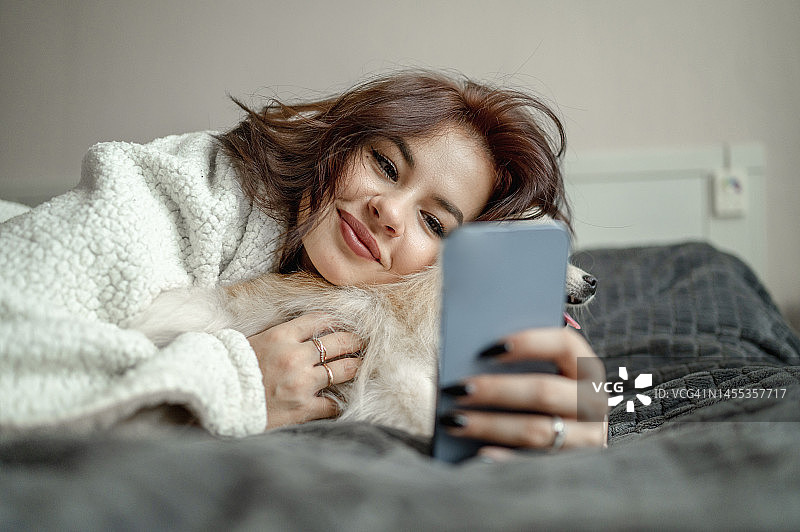 微笑的女人在家抚摸博美犬并使用智能手机图片素材
