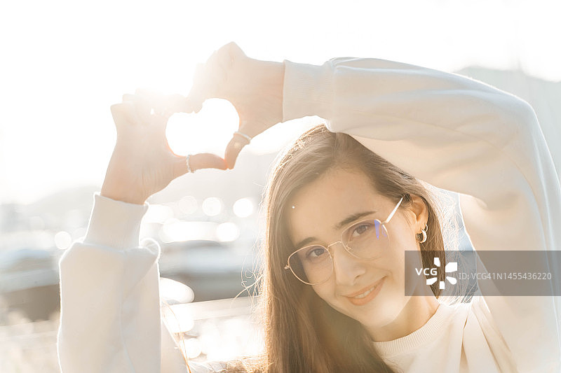 身穿白色运动衫的美丽年轻女子肖像。在天空背景上手比成心形。阳光明媚的一天，白色。图片素材