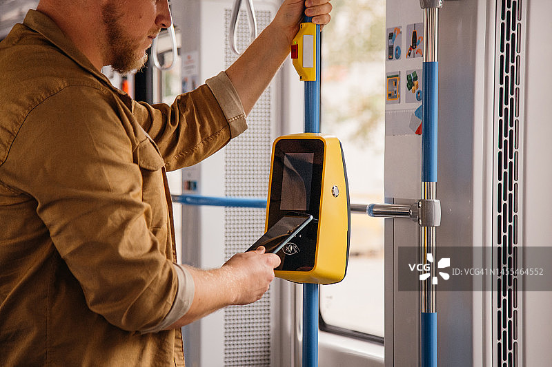 非接触式支付：年轻男人在公共交通工具上进行支付。男人用手机支付乘坐电车费用。男性通过数字钱包购买车票。特写图片素材