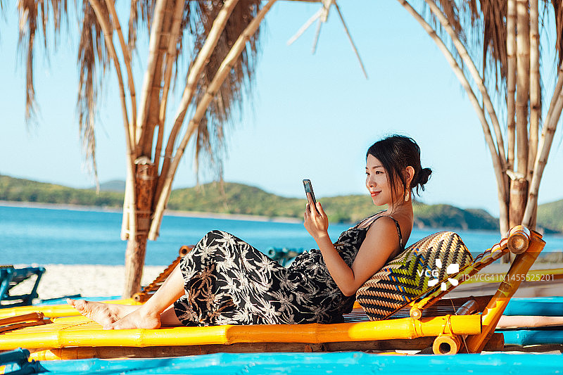 躺在海滩长椅上使用智能手机的年轻亚洲女性图片素材