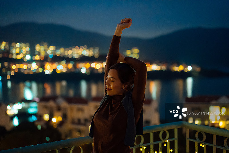 年轻漂亮的亚洲女性在阳台上放松身心，伸展双臂，感受焕然一新，背景是城市夜景图片素材