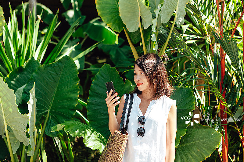 年轻亚洲女人在户外热带植物前使用智能手机图片素材