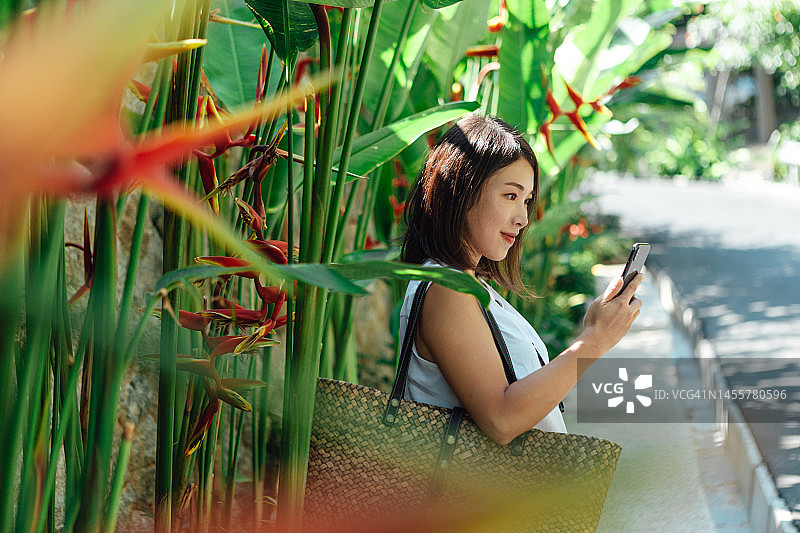 年轻亚洲女性在户外热带植物背景下使用智能手机的侧视图图片素材