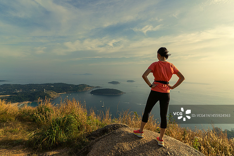 一名女子身穿登山服站在岩石上，眺望普吉镇、山脉、大海和停泊在黑石崖下的船只，泰国普吉府图片素材