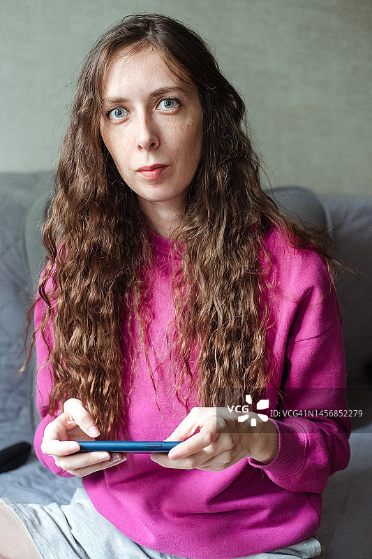 一位穿着亮粉色运动衫，手拿电话，坐在家中灰色沙发上的年轻长发卷发女孩的肖像图片素材