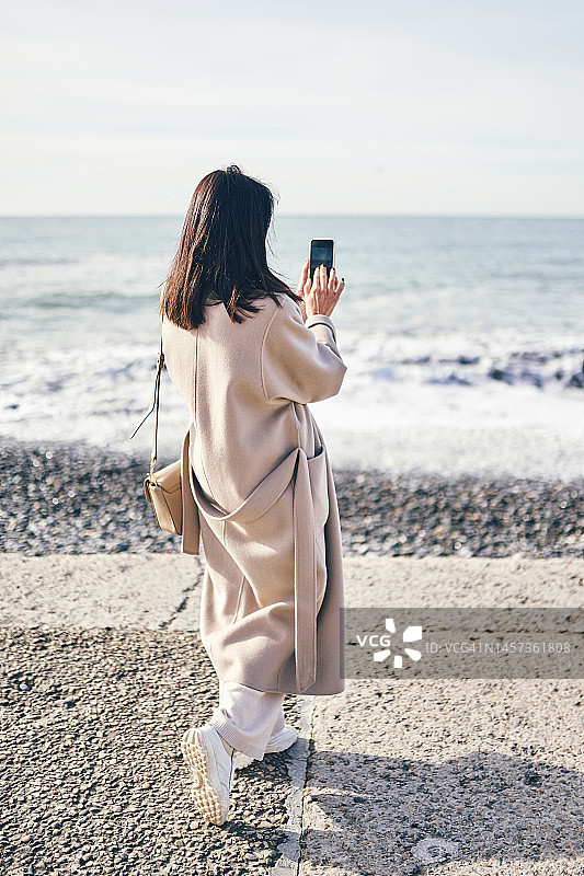 一位身穿外套、留着深色头发的女子站在海边，拿着手机拍照图片素材