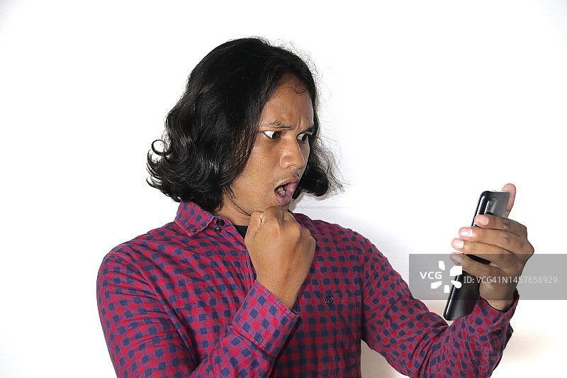 手持智能手机，打字、与社交网络中的朋友聊天、玩在线游戏的，面部表情惊讶的年轻亚洲男人，与白墙隔离图片素材