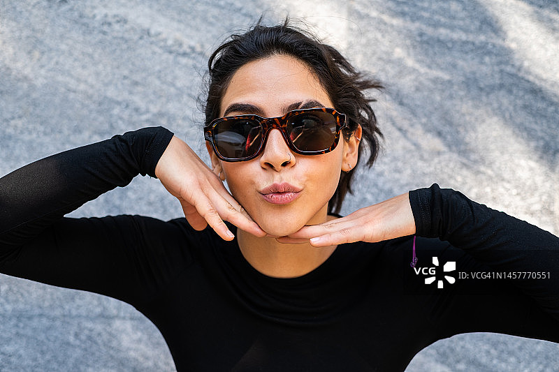 戴着墨镜和鼻环的拉丁女性看着镜头，用手勾勒脸庞，灰色大理石背景半身照图片素材