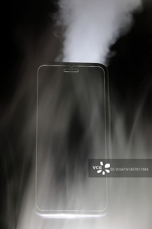 黑色背景中具有白色蒸汽、烟雾和雾气效果的智能手机。带有屏幕钢化玻璃盖的移动设备，提供安全保护。图片素材