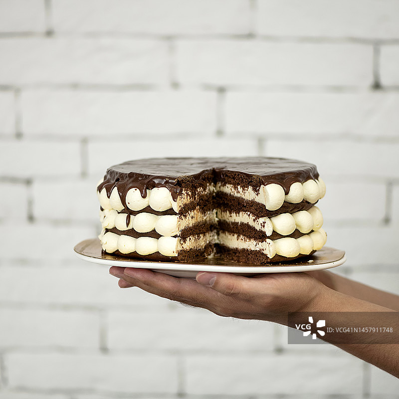 手中盘子里的切块蛋糕：家庭庆祝的奶油巧克力甜点，白色砖墙背景，柔焦，侧视图，剖视图图片素材