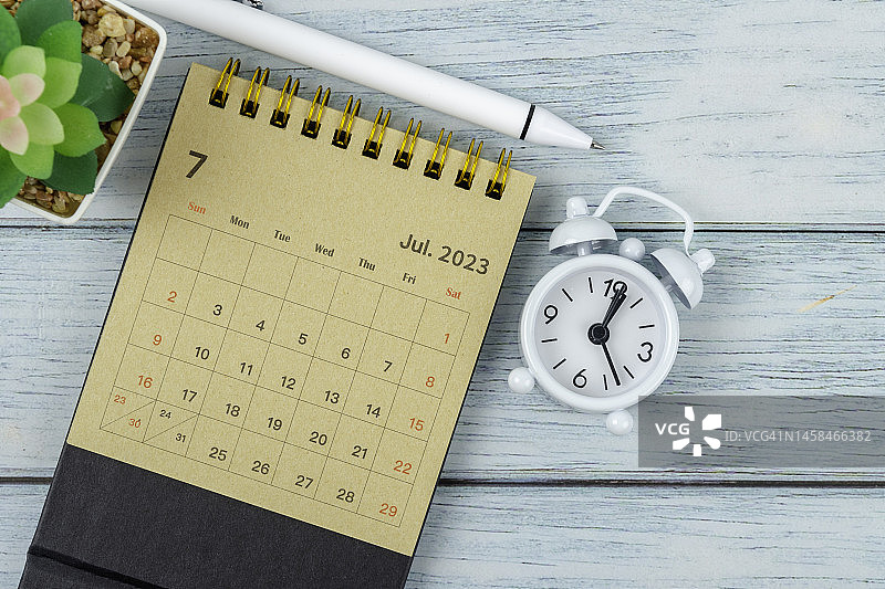 2023年7月桌面日历：木桌背景上的闹钟和白色笔图片素材
