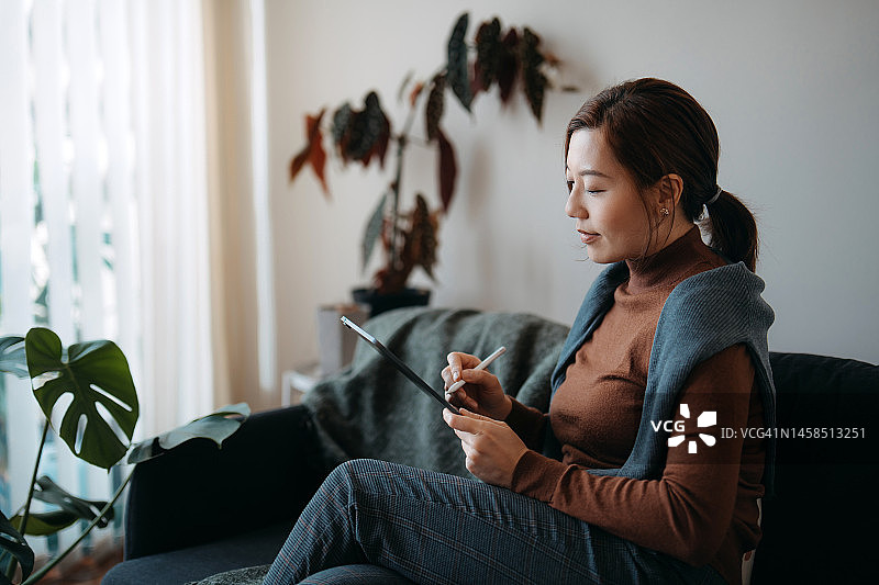 在家中客厅沙发上使用平板电脑的年轻漂亮亚洲女性，背景是室内植物。居家办公。自由职业者。灵活的工作与生活平衡。日常生活中科技的应用图片素材