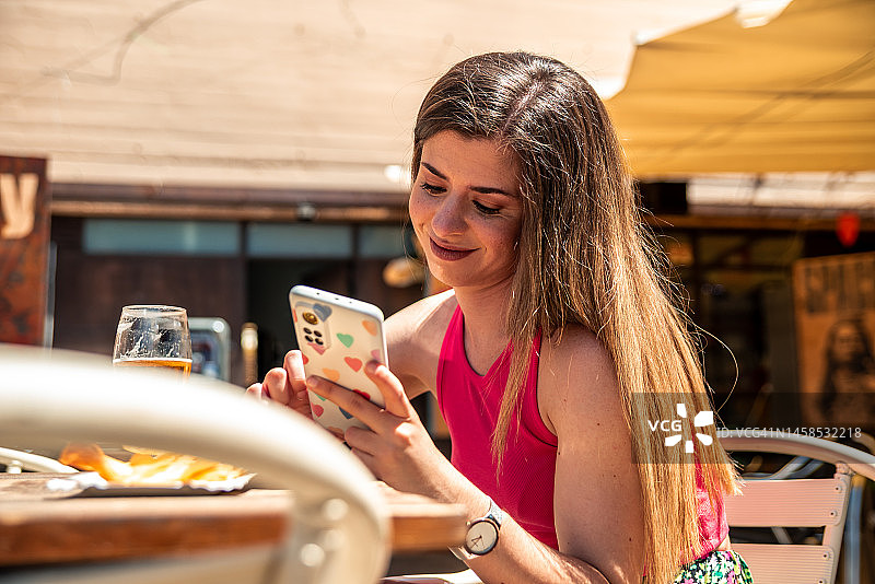 年轻金发女孩在酒吧露台上喝啤酒、吃薯条，同时使用手机图片素材