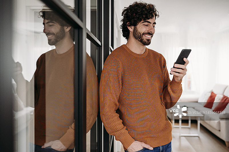 微笑的年轻人在家站在玻璃门旁使用手机图片素材