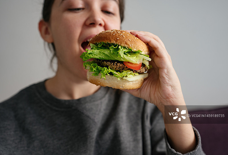 坐在沙发上吃着一个夹着肉、番茄和生菜的大汉堡的女人，快餐概念图片素材