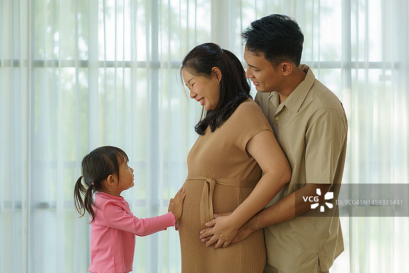 亚洲女儿触摸孕妇的肚子，父亲拥抱妻子，象征爱与家庭的联系图片素材