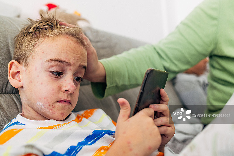 母亲给儿子涂抹药膏，缓解水痘引起的瘙痒，儿子在玩手机游戏图片素材