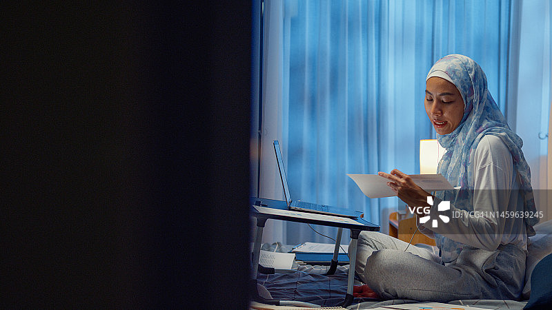 快乐的年轻穆斯林女人坐在床上，在折叠笔记本电脑桌上使用电脑，在家工作图片素材