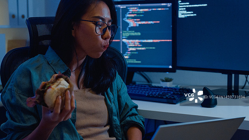 年轻亚洲女性软件开发人员在家远程工作，吃汉堡包，用电脑编写代码图片素材
