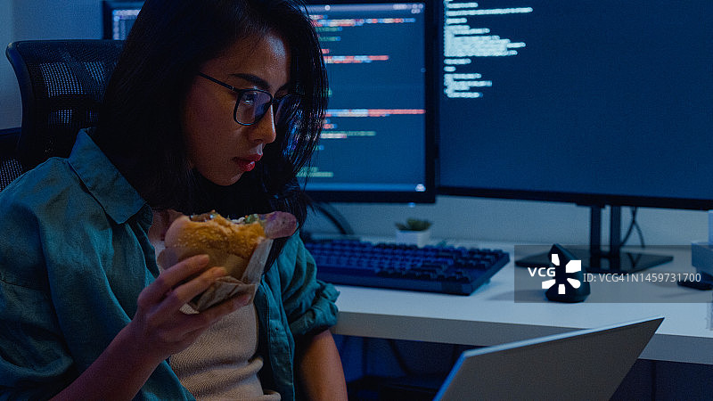 年轻亚洲女性软件开发人员在家远程工作，吃汉堡包，用电脑编写代码图片素材
