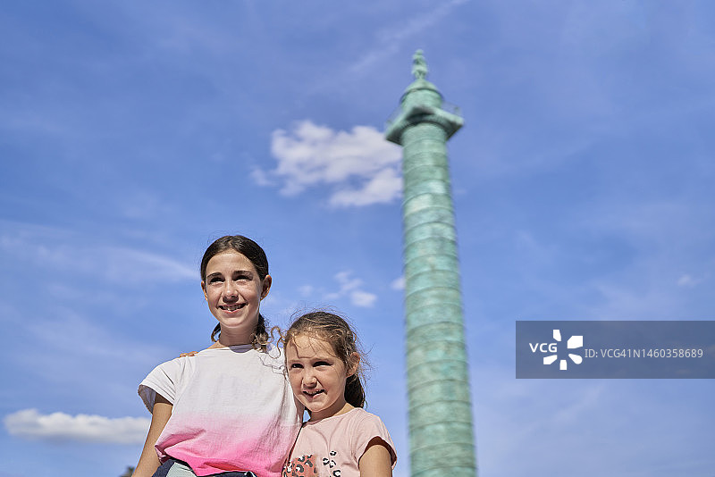 在巴黎旺多姆广场绿色圆柱背景下，两个女孩微笑拥抱看向镜头图片素材