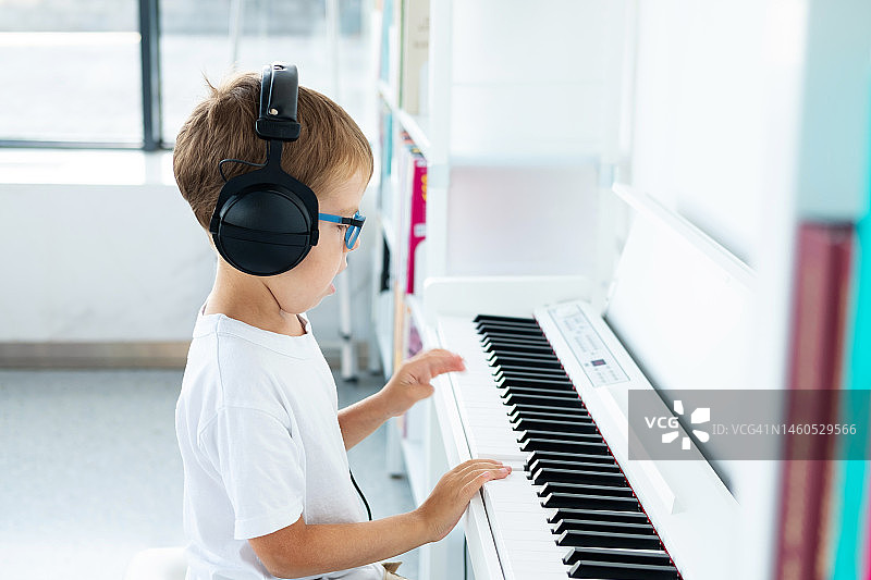 戴眼镜、大耳机、穿白色T恤的小男孩在音乐课上弹奏白色数码钢琴图片素材